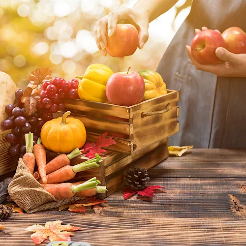 A hand reaches toward apples while an autumn harvest of fruits and vegetables sits in wooden crates on a rustic table.