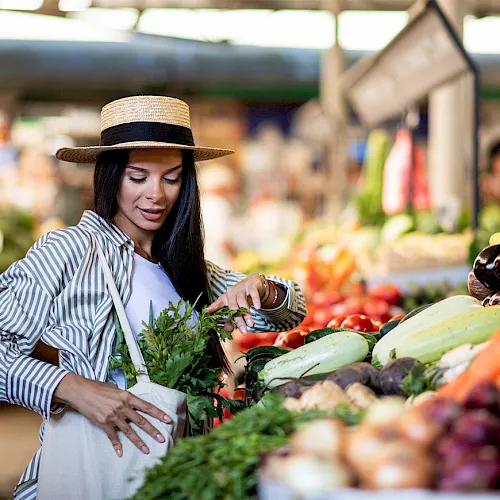 A woman in a straw hat arranges fresh herbs at a colorful outdoor market stall.