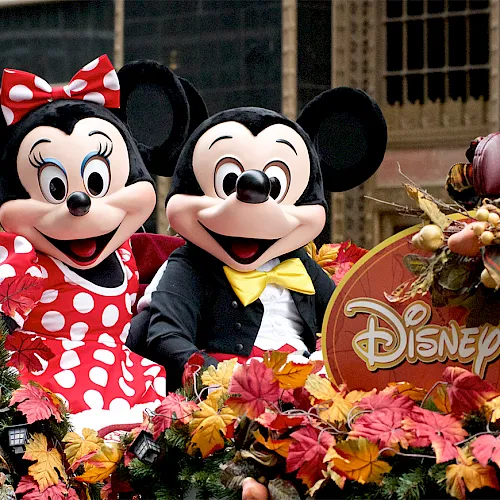 Minnie and Mickey Mouse pose at a Disney fall-themed display with colorful autumn leaves and decorations.