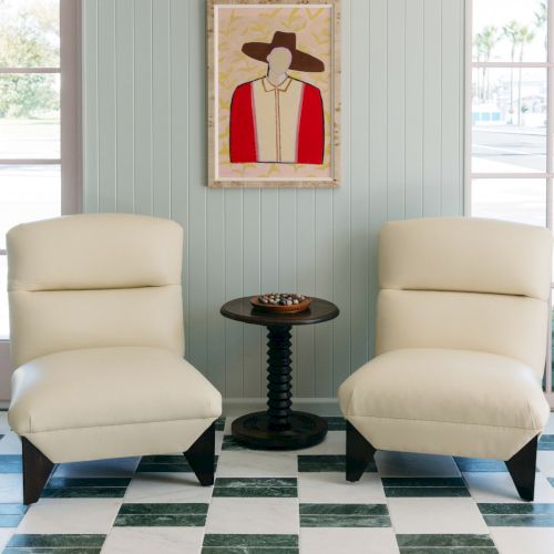 A bright lounge corner with two cream chairs, a small round table, and a framed portrait painting on striped walls.