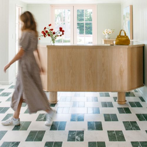 A blurred woman in a light dress walks past a wooden reception counter with flowers and a yellow bag in a bright room.