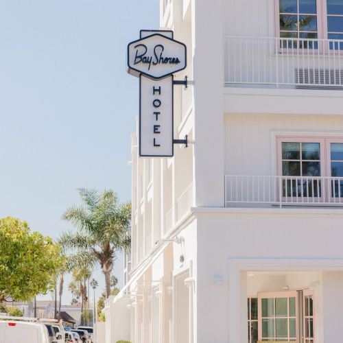 A street view of a white building with a &ldquo;Bay Shores&rdquo; hotel sign and roadside greenery and parked vans in the background.