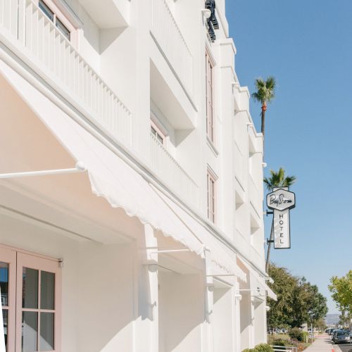 White hotel building with awnings and windows, plus a Bay Shores Hotel sign and palm trees under a clear blue sky.