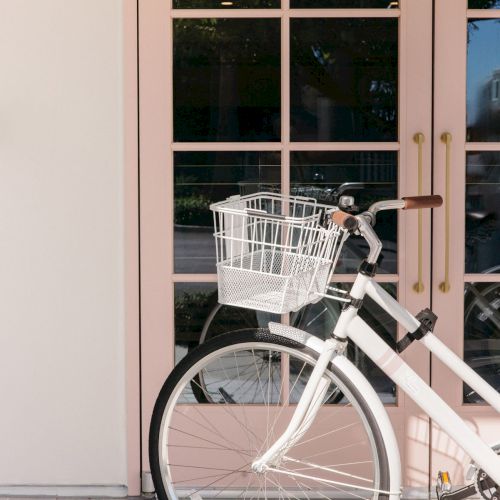 A white bicycle with a front wire basket is parked by a building&rsquo;s large window in warm daylight.