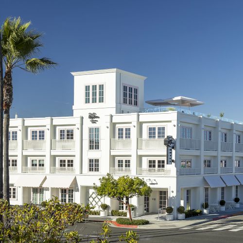 A sunny white multi-story building with palm trees and outdoor umbrellas in front under a clear blue sky.