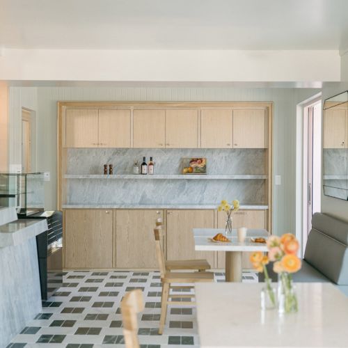 A bright modern kitchen with wood cabinets, marble counters, pendant lights, and a small dining area with flowers.