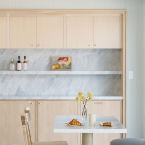 A modern kitchen with wood cabinetry, a marble backsplash, wine bottles on a shelf, and croissants on a table with flowers.