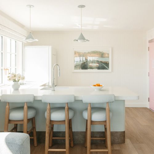 A bright modern kitchen with a long counter, three stools, pendant lights, a sink, and framed wall art.