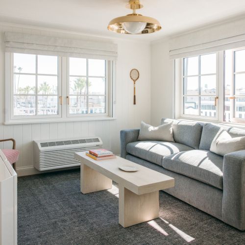 A bright living room with gray sofa, coffee table, windows with blinds, a wall art print, and a ceiling light fixture overhead.