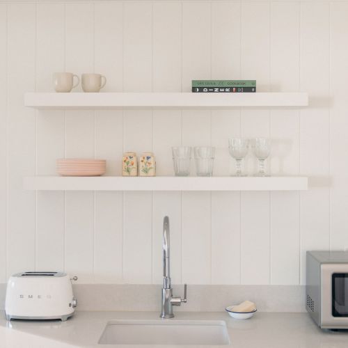 A bright kitchen with floating shelves, mugs and glasses above the counter, plus a toaster, sink, and microwave.