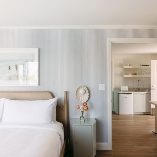 A bright bedroom with striped wallpaper, a bed and framed beach photo, opening to a small kitchenette with shelves, sink, and a fridge.