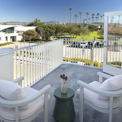 Sunny resort balcony overlooking palm trees, green lawn, parked cars, and distant mountains beyond a modern building.