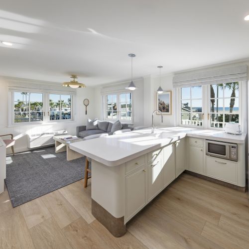 Bright modern coastal kitchen with a white island, sink, stove and microwave, plus living area and ocean-view windows under ceiling lights.