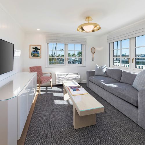 Bright living room with a gray sofa, TV, coffee table, and large windows with blinds overlooking palm trees.