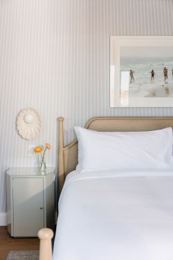 A bright bedroom corner with a wooden bed, white pillows, a bedside cabinet, a glass vase with a yellow flower, and beach wall art.