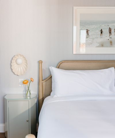 A bright bedroom corner with a wooden bed, white pillows, a bedside cabinet, a glass vase with a yellow flower, and beach wall art.