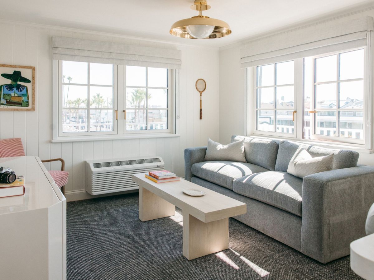 A bright living room with a gray sofa, coffee table, framed art, large windows, and a gold ceiling light.