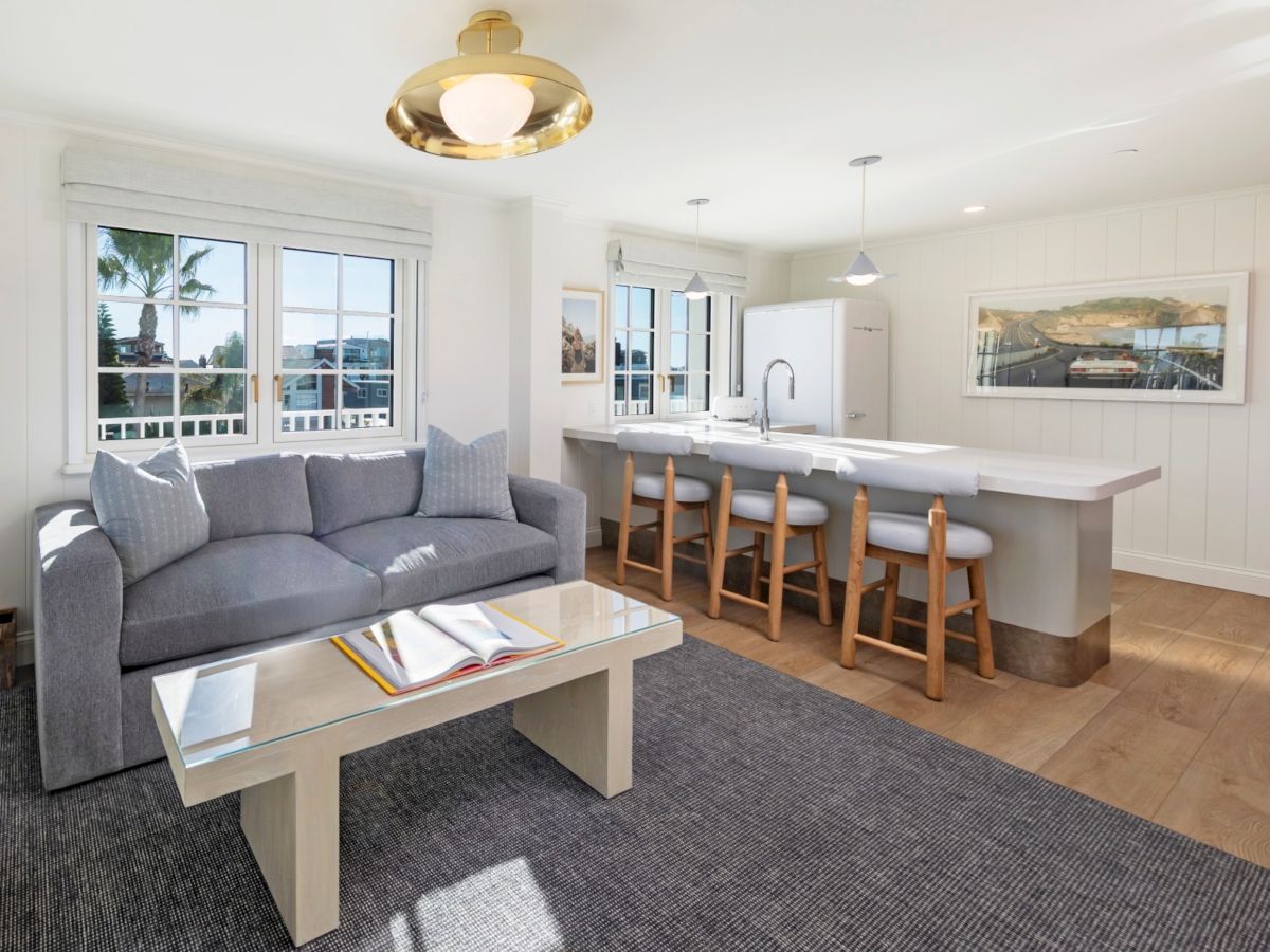 A bright living room with a gray sofa, coffee table, kitchen island with stools, pendant lights, large windows, and wall art.