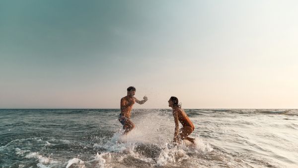 A couple splashing and laughing in the ocean waves at the beach, with spray bursting around them under a clear sky.