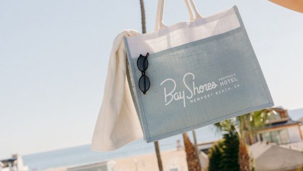 A person holds a Bay Shores Hotel tote bag above a sunny street with palm trees in the background.