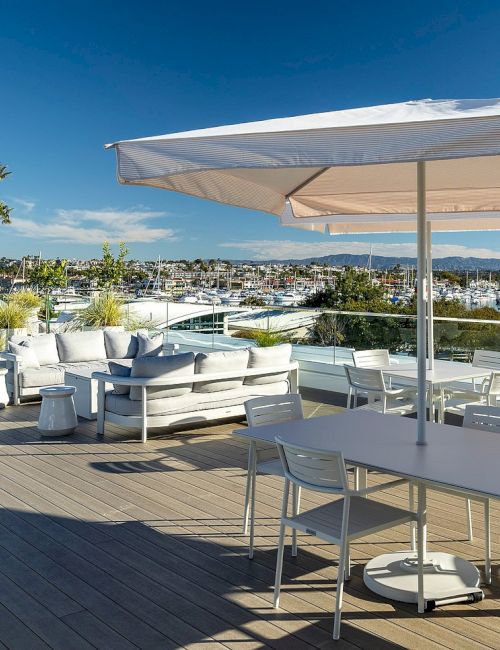 An outdoor patio with white cushioned seating, a large striped umbrella, and dining table overlooking a sunny marina.