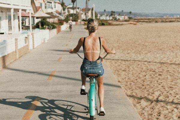 A person rides a blue bicycle along a seaside path beside sand and buildings under a clear blue sky.